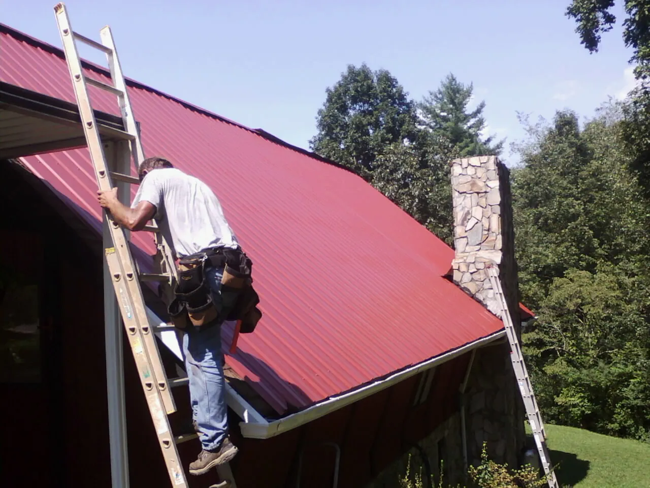 Roofer on a ladder beside a red metal roof and stone chimney — Soffit & Fascia Repair by Roofing Brothers in Nada